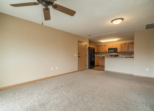 a spacious living room with a ceiling fan, next to a kitchen with wooden cabinets and a tall bar top counter