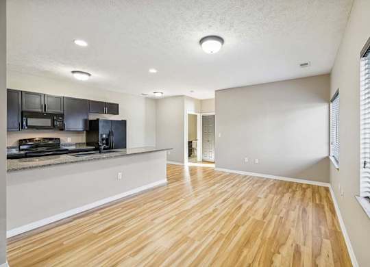 A large living room with wooden floors and a large kitchen island. Black appliances are in the kitchen. There is a hallway in the background.