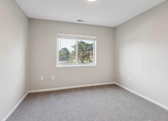 Bright bedroom with carpet flooring and a wide window overlooking neighboring buildings, providing natural light and a clean, neutral interior.