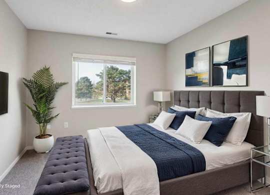 Virtually staged bright apartment bedroom with large window, neutral walls, upholstered bed, and modern furnishings with soft natural light.