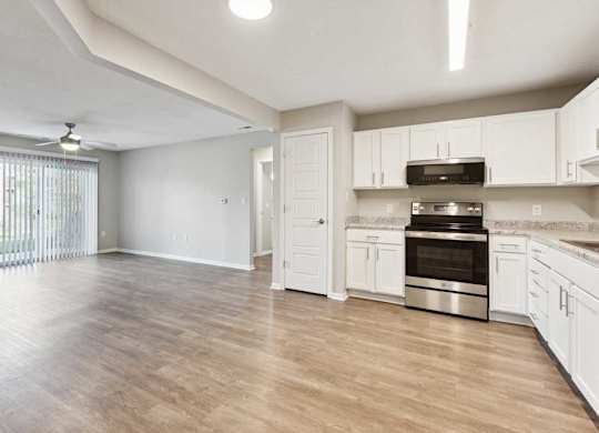 Open-concept apartment interior featuring kitchen with white cabinets and stainless steel oven, flowing into a spacious living area with wood-style flooring.