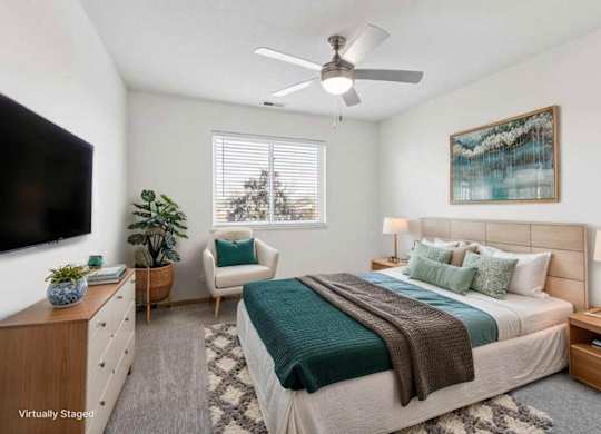 Virtually staged apartment bedroom with ceiling fan, large windows, neutral finishes, and cozy seating area beside the bed.