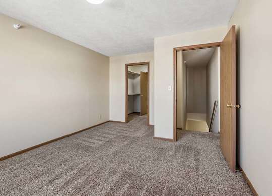 Spacious apartment bedroom with neutral carpet, beige walls, wood-trimmed doors, and an open layout leading to the hallway and closet area.