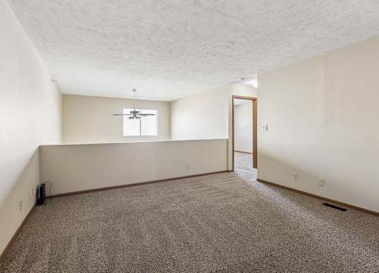 Spacious loft area in an apartment with neutral carpet, a half wall overlooking the lower level, a window for natural light, and an open doorway leading to the hallway.