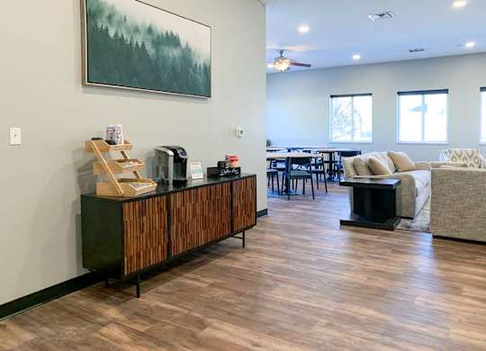 a clubhouse kitchen and tv lounge with wooden floors and furniture, a painting on the wall, and a wooden cabinet with items on top