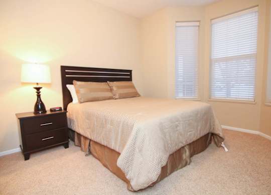 A bedroom with a bed, nightstand, and lamp, plus bay windows