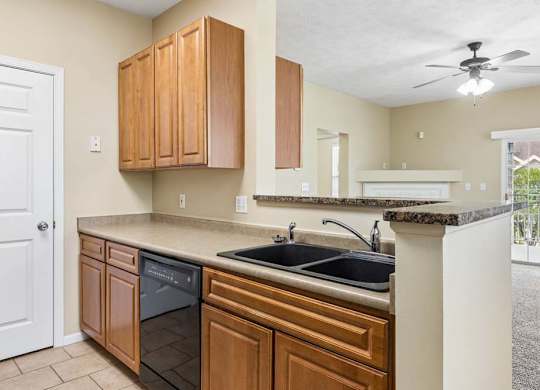 Apartment kitchen with wood cabinetry, double sink, black dishwasher, breakfast bar, and view into living room.