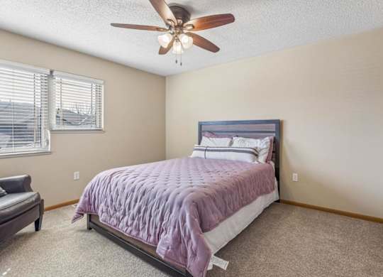 Bright apartment bedroom with neutral carpet, large corner windows, ceiling fan, and a queen-size bed, offering a comfortable and airy interior.