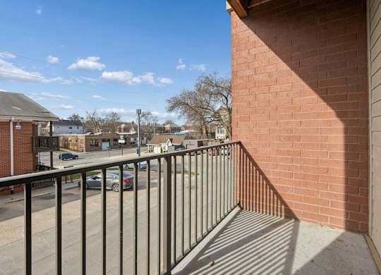 Private apartment balcony with a black metal railing, brick exterior, and neighborhood views under a clear blue sky.