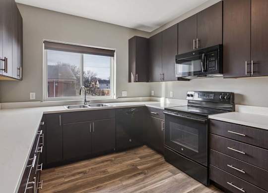Modern apartment kitchen with dark wood cabinetry, black appliances, white countertops, and a window over the sink.