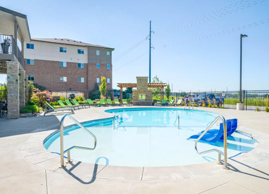 a swimming pool with blue chairs in shallow water and an apartment building in the background