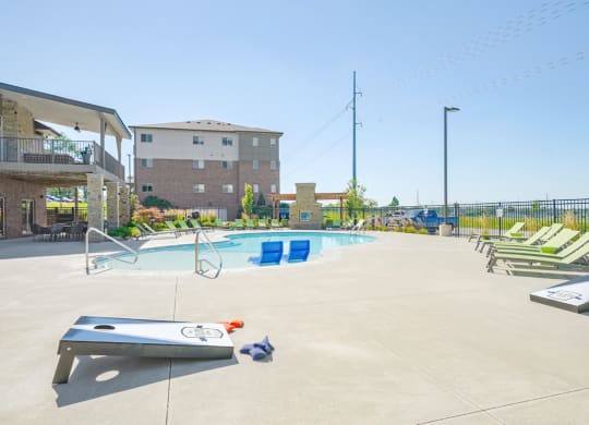a pool with green lounge chairs and a cornhole game in front of a building