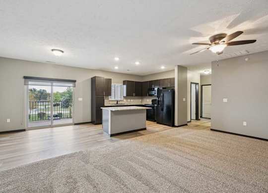 A spacious, carpeted living room open to a modern kitchen with hardwood flooring and dark wooden cabients.