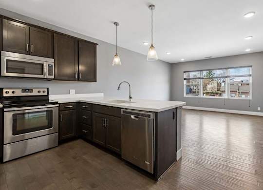 Dark cabinetry and a white peninsula define this open apartment kitchen, flowing into a living area with wide windows and wood-look floors.