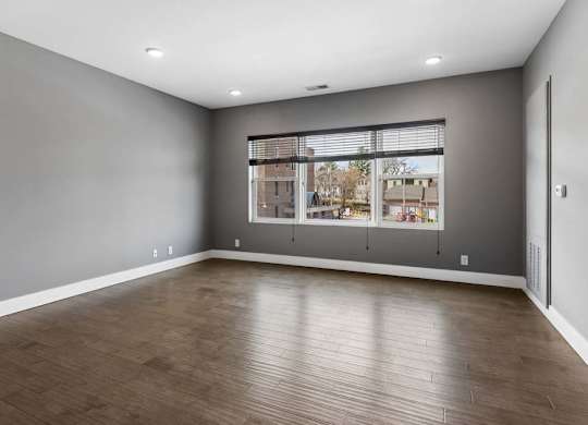 Expansive apartment living room features wood-look flooring, gray walls, and a broad window overlooking the surrounding community.