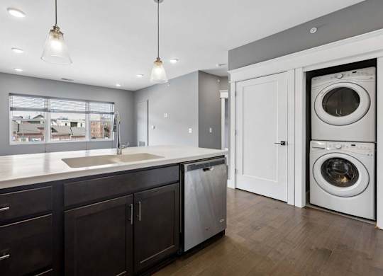 Stacked in-home laundry sits just off the kitchen, beside a dark cabinet island with a deep sink, dishwasher, and pendant lights.