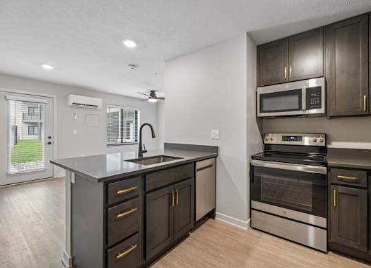 Modern apartment kitchen with dark cabinetry, stainless steel appliances, a center island with storage, and an open layout connected to the living area.