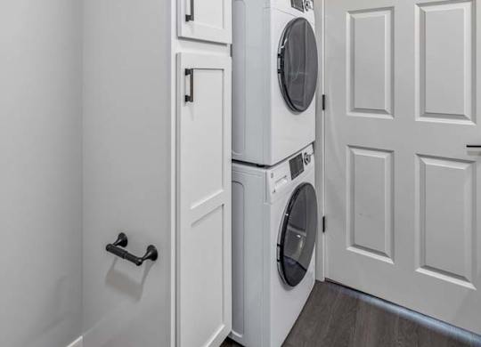Apartment laundry area with stacked washer and dryer, storage cabinetry, and wood-style flooring.