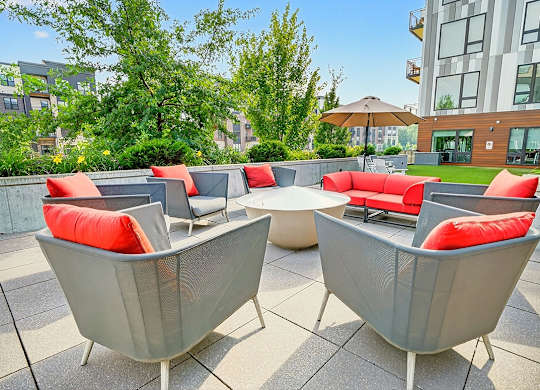 A fire pit surrounded by gray lounge chairs and red-cushions on a patio with nearby greenery.