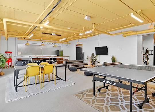 Bright game room with yellow ceiling, featuring a ping pong table, foosball table, and a dining area with four yellow chairs.