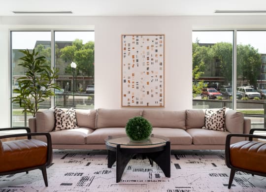 Community lounging space with large beige couch in between two large windows. Two chair sit opposite of the couch with a small coffee table at the center.