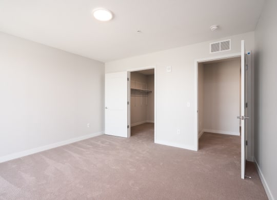 large bedroom with beige carpet and white walls. Two open doors, one leading to the walk in closet and the other leads to the rest of the apartment.