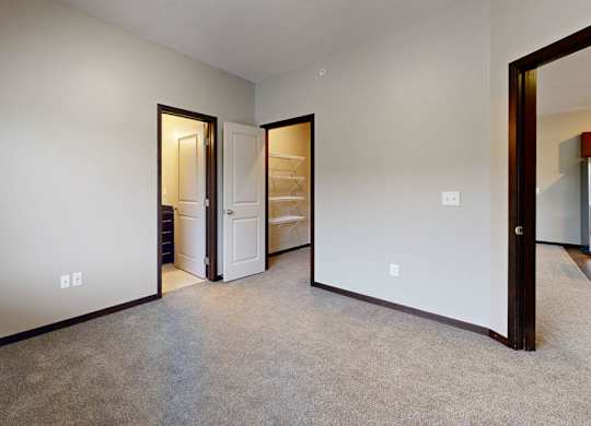 A room with a carpeted floor and a white wall with 2 doorways leading to bathroom and closet
