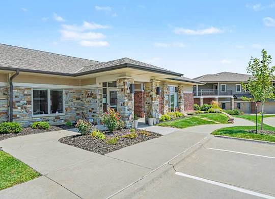 Front view of apartment leasing office with glass double doors, stone and brick exterior, manicured lawn, small tree, and parking spaces in foreground.