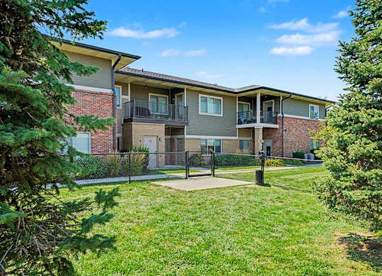 Two-story apartment building with brick and siding exterior, private balconies, and a fenced green lawn area with trees and open sky.