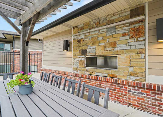 Outdoor apartment community patio with pergola, long dining table and chairs, and a stone accent wall with built-in fireplace and modern wall lighting.