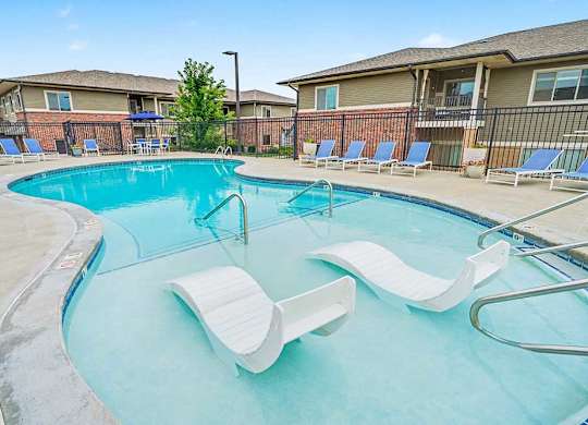 Resort-style apartment pool with curved design, shallow sun shelf and in-water lounge chairs, surrounded by deck seating and fenced residential buildings.