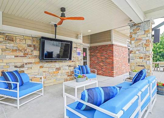 Covered poolside lounge at an apartment community with blue cushioned seating, ceiling fans, stone columns, and views of the outdoor swimming pool and sundeck.