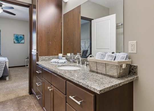 Staged apartment bathroom vanity with speckled countertop, dark wood cabinets, large mirror, and tile flooring.