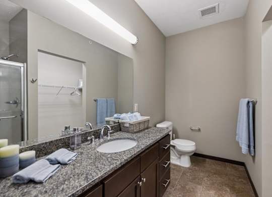 Apartment bathroom with speckled vanity, dark wood cabinetry, large mirror, tile floor, and walk-in closet access.