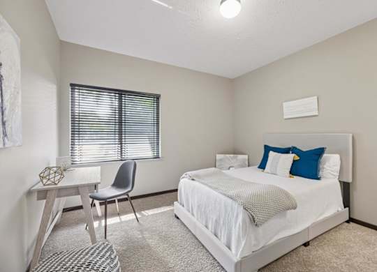 Bright staged apartment bedroom with carpet, upholstered bed, desk, window with blinds, and neutral finishes.