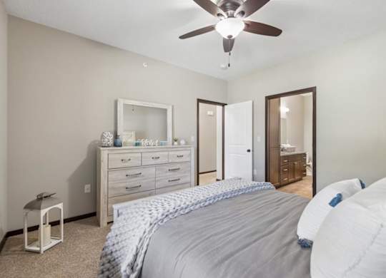 Staged apartment bedroom with ceiling fan, dresser, carpet, and direct access to the bathroom.