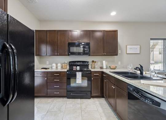 Staged apartment kitchen with dark wood cabinets, black appliances, speckled counters, and tile flooring.