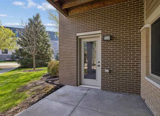 Covered apartment patio with brick wall, glass door, concrete slab, and landscaped grounds.