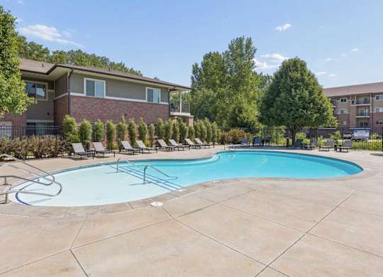 Outdoor apartment community swimming pool with lounge chairs, wide concrete deck, mature trees, and residential buildings.