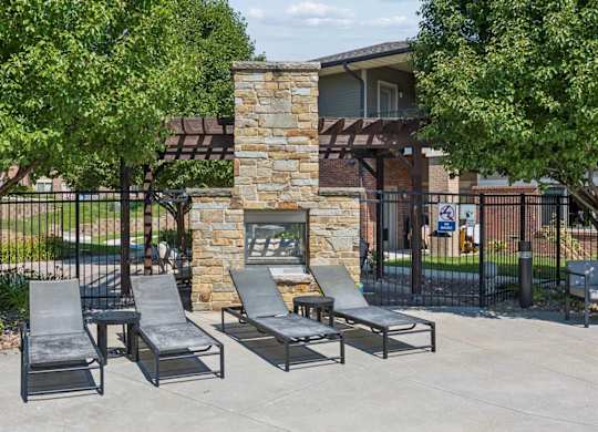Poolside lounge area at Villas of Omaha at Butler Ridge with chaise seating, a stone fireplace, pergola, and mature trees.