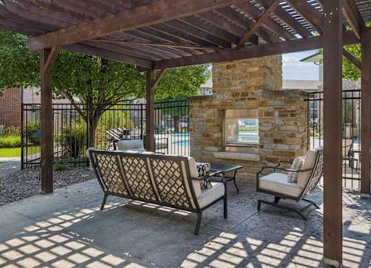 Pergola-covered outdoor lounge at Villas of Omaha at Butler Ridge with cushioned seating and a stone fireplace beside the pool.
