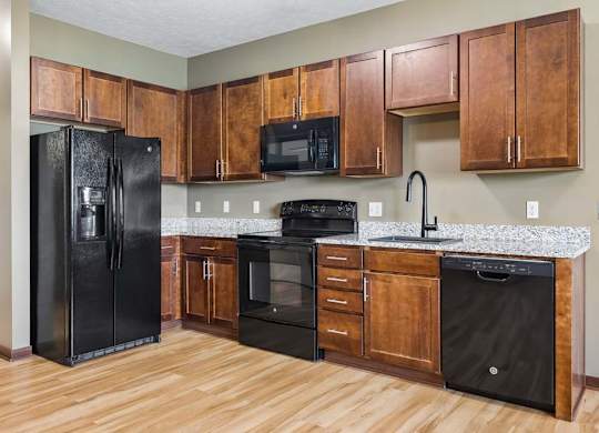 Apartment kitchen with warm wood cabinetry, speckled countertops, black appliances, wood-look flooring, and a tall curved faucet.