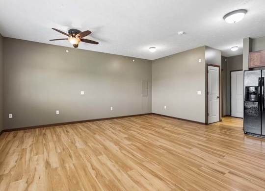 Bright, empty apartment living area with wood-look flooring, neutral walls, ceiling fan, and an open view into the kitchen.