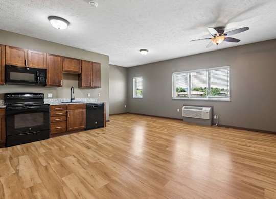 Open-concept apartment kitchen and living area with wood-look flooring, dark appliances, wood cabinetry, ceiling fan, and large windows.