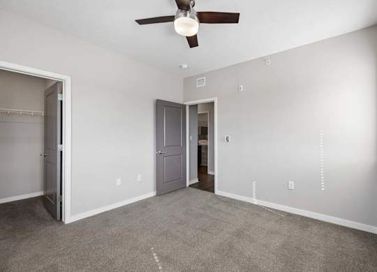Spacious apartment bedroom with carpeted flooring, a ceiling fan, a walk-in closet, and a window with blinds providing natural light.