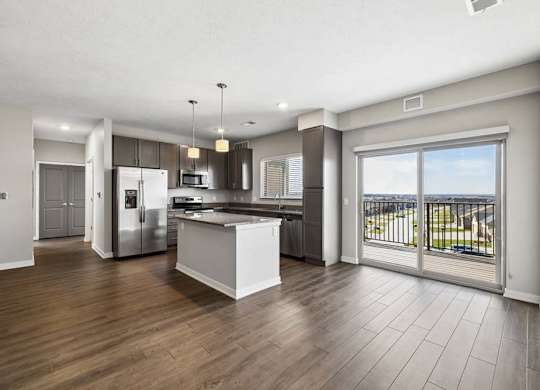 Spacious open-concept apartment living area with wood-style flooring, a modern kitchen featuring a center island, pendant lighting, stainless steel appliances, and a large sliding glass door leading to a private balcony.