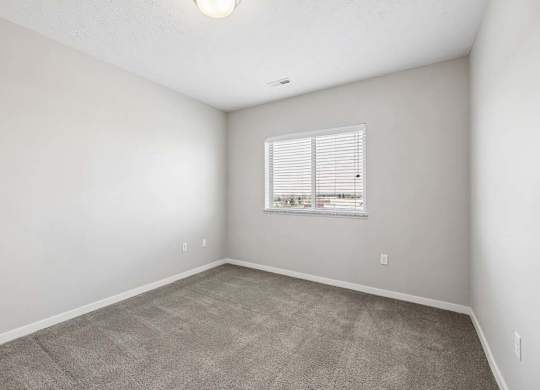 Bright apartment bedroom with carpeted flooring, neutral walls, and a window with blinds providing natural light.
