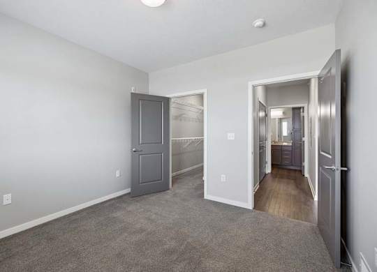 Bright apartment bedroom with carpeted flooring, an open closet with sliding doors, gray interior doors, and access to the adjoining hallway.