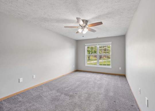 Bright apartment bedroom with soft carpeting, a ceiling fan, and a large window providing natural light.