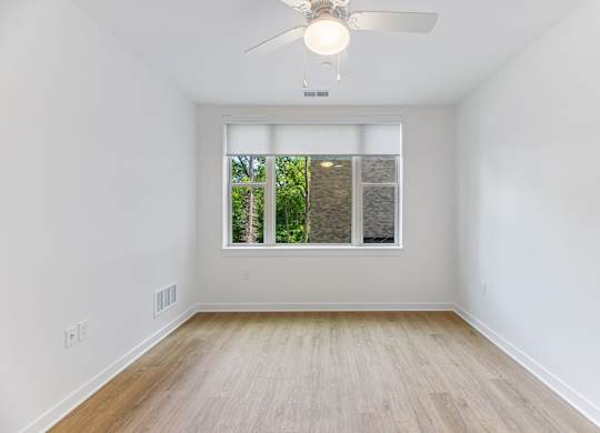 A room with a ceiling fan and a window with a view of trees.